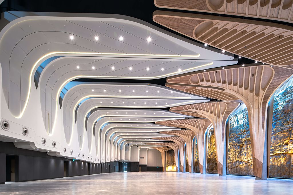 Futuristic hallway with curved white ceiling panels and wooden Y-shaped supports, illuminated by ambient lights along the arches.