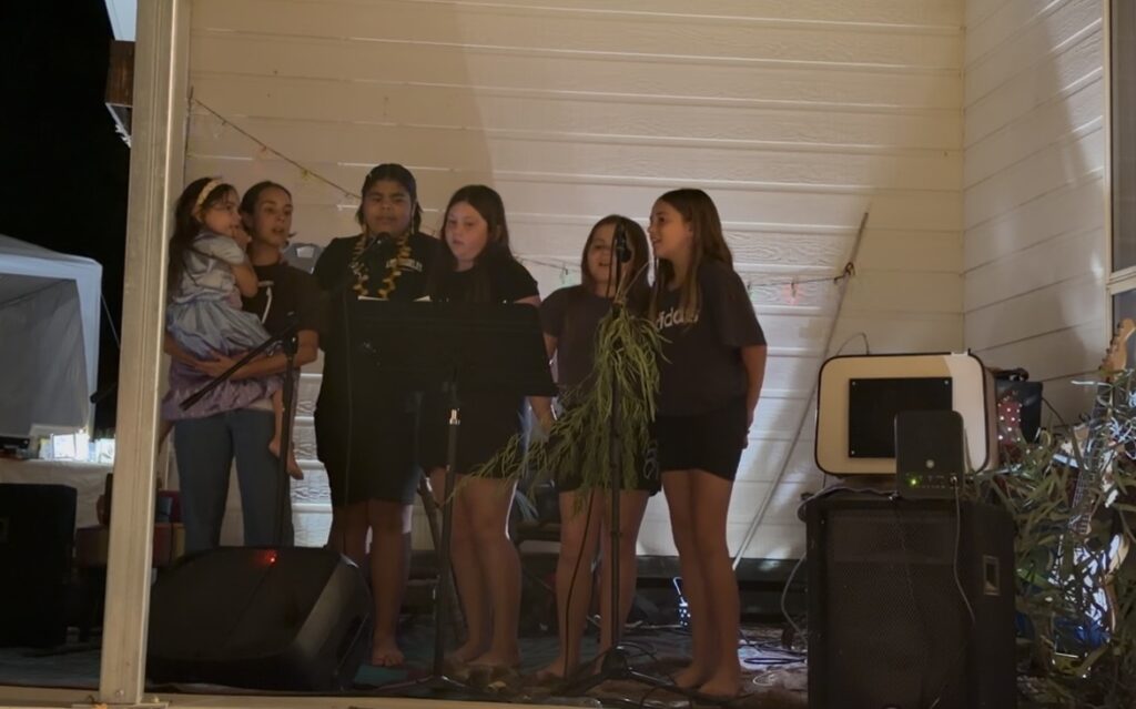 Group of young girls singing into microphones on an outdoor stage at night, one girl holding a small child nearby.