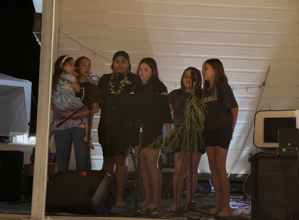 Group of young girls singing into microphones on an outdoor stage at night, one girl holding a small child nearby.