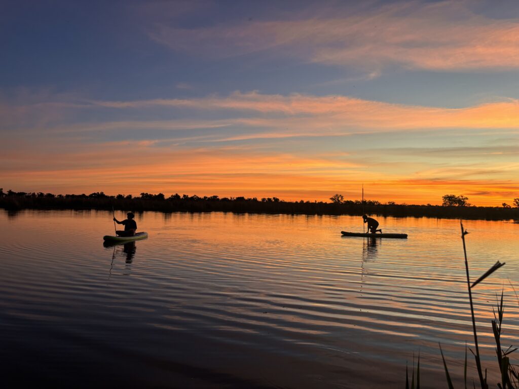 Two people paddleboarding on a calm lake at sunset, silhouetted against an orange-pink sky with a distant tree line.