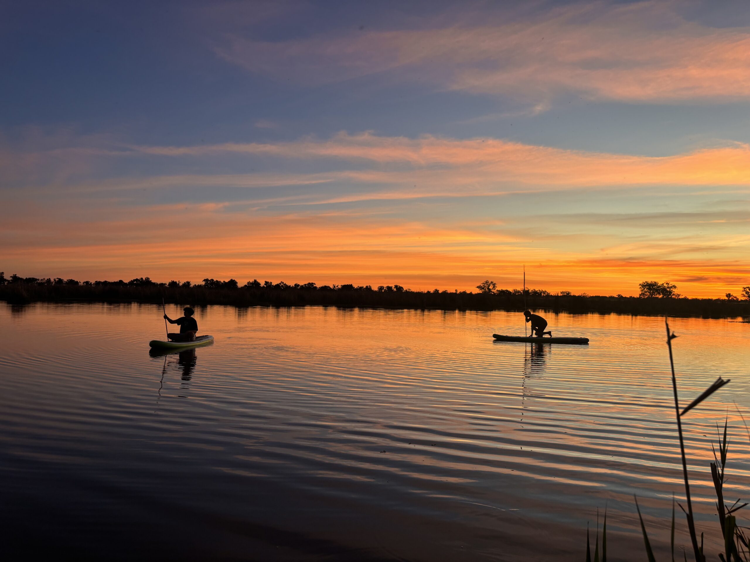 Two people paddleboarding on a calm lake at sunset, silhouetted against an orange-pink sky with a distant tree line.